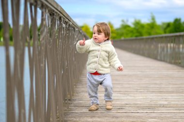 Kabarık ceketli, eşofmanlı küçük çocuk tahta bir köprüde yürüyor..