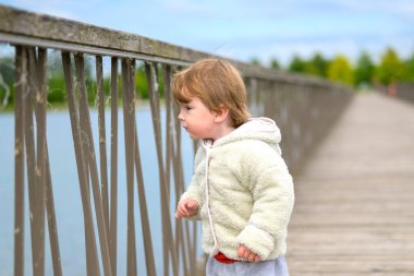 Kabarık kapüşonlu ceketli küçük bebek tahta bir köprüde duruyor, merakla korkuluklardan suya doğru bakıyor..