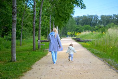 Sarışın bir kadın ve yeni yürümeye başlayan bir çocuk, ağaçlar ve uzun otlarla kaplı geniş bir park yolunda yan yana yürüyorlar..