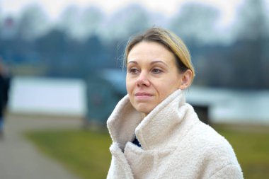 Yüksek yakalı, krem rengi ceketli bir kadın serin bir günde dışarıda duruyor..