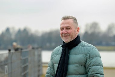 Yeşil kabarık ceketli ve siyah eşarplı bir adam sakin bir gölün yanında metal bir çitin yanında dururken sıcak bir şekilde gülümsüyor..