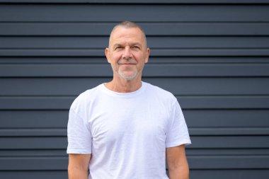 Kısa gri saçlı, sakallı, modern, koyu gri duvarın önünde duran kendinden emin, olgun bir adam..