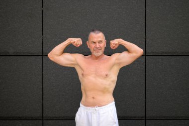 Üstsüz kıdemli bir adam güçlü bir pozda iki kolunu sallarken hafifçe yana bakıyor..