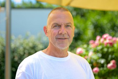 Beyaz tişört giyen olgun bir adamın portresi. Çiçek açan bir bahçenin önünde dikiliyor..