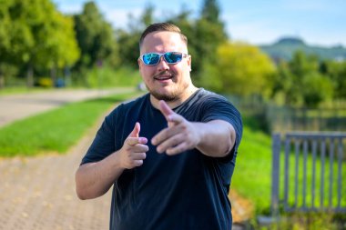 Yansıtıcı güneş gözlüğü takan genç bir adam ve siyah bir tişört şakacı bir şekilde kamerayı işaret ediyor..