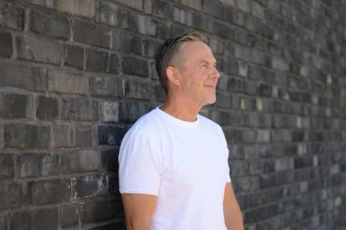 Beyaz tişörtlü, olgun bir adamın, desenli tuğla bir duvarın karşısında gelişigüzel dikilmesinin yan görüntüsü..