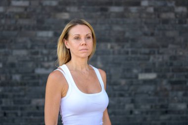 Koyu tuğlalı bir duvarın önünde duran, beyaz kolsuz bir bluz giyen ve sakin, ciddi bir ifadeyle ileriye bakan kendine güvenen sarışın kadın..