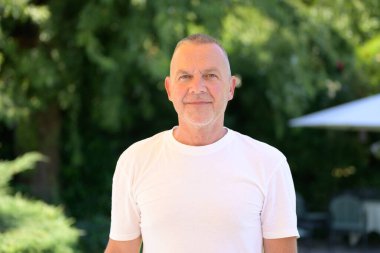 Sakin, beyaz tişörtlü yaşlı bir adam güneşli yeşil bir bahçede duruyor..