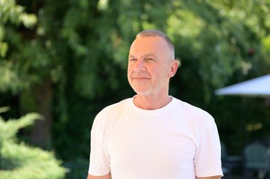 Beyaz tişörtlü düşünceli bir son sınıf öğrencisi güneşli bir bahçede dikilirken yüzünü çeviriyor..