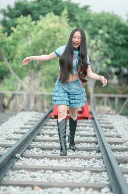 Canlı genç bir kadın trenin raylarında mutlu bir şekilde yürüyor, trend kıyafetleri içinde, etrafı yemyeşil, özgürlüğün ve maceranın somutlaştırılmış bir şekilde.