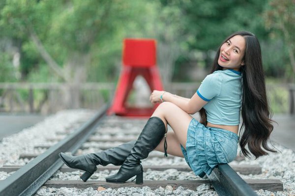 Веселая молодая женщина позирует на железнодорожных путях, одетая в кукурузный топ и джинсовую юбку, излучая радость на ярком зеленом фоне на открытом воздухе.