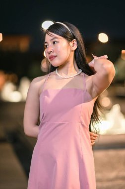 Pembe elbiseli genç bir kadın gece zarif bir şekilde dışarıda duruyor. Etrafı güzel bokeh ışıkları ve su özellikleriyle çevrili. İnce bir güzellik ve sofistike anı yakalıyor..