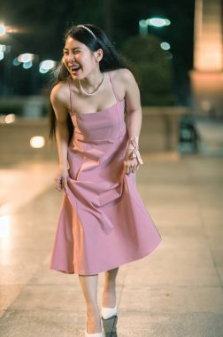 Genç bir kadın, gece gezintisi sırasında pembe bir elbise giyerken neşeyle gülüyor, şehir sahnesine ışıl ışıl enerjisiyle canlı bir atmosfer ekliyor..