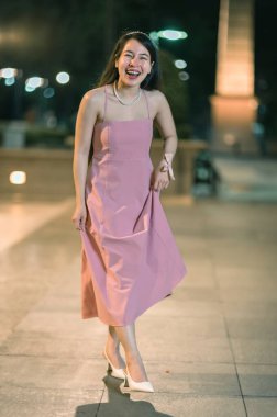 A cheerful young woman embodies joy in a stylish pink dress while walking through an urban setting at night, illuminated by warm street lights. Perfect for fashion and lifestyle themes. clipart