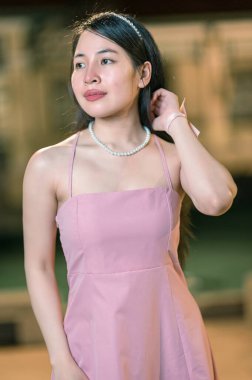 Şık pembe elbiseli ve inci kolyeli genç bir kadın zarifçe poz veriyor, zarafet ve duruş sergiliyor. Hafifçe aydınlatılmış arka plan güzelliğini ve çekiciliğini arttırır..