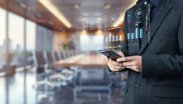 Modern bir konferans odasında sergilenen dijital verileri analiz etmek için akıllı telefon kullanan bir iş adamı, kurumsal analiz ve stratejiyi sembolize ediyor..