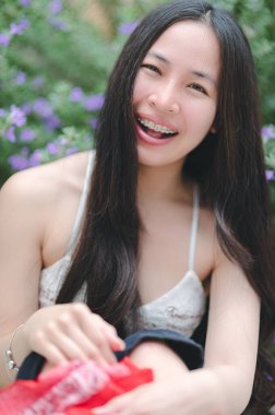 Uzun saçlı neşeli genç bir kadın çiçek açan çiçeklerle çevrili, gür bir bahçede parlak bir şekilde gülümsüyor. Neşeli ifadesi bir anlık mutluluk ve kaygısız ruhu yakalar..