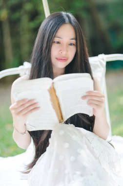 Serene young woman sits outdoors, reading a book in a soft white dress. Surrounded by nature and sunlight, she embodies tranquility and contemplation, enjoying a peaceful moment. clipart