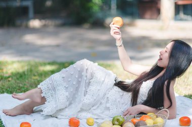 Neşeli genç bir kadın piknik battaniyesinde dinleniyor, açık havada taze meyvenin tadını çıkarıyor. Bu canlı sahne doğada mükemmel bir günü yakalar..