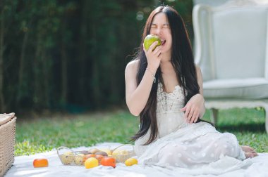 Neşeli genç bir kadın açık havada taze meyveyle çevriliyken yeşil bir elmayı ısırıyor. Huzurlu atmosfer, doğal güzelliği ve sağlıklı yaşamı vurgular..