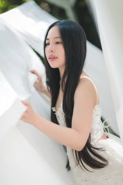 Uzun saçlı genç bir kadın, beyaz kumaşın arasında zarif bir şekilde poz verir. Yumuşak güneş ışığında dingin bir güzelliği yakalar. Sahne huzuru ve zarafeti çağrıştırıyor..