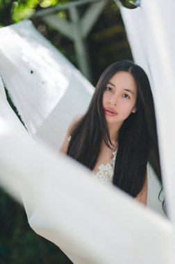 Uzun siyah saçlı, beyaz kumaşa batmış genç bir kadının portresi. Sakin atmosfer ve yumuşak güneş ışığı sakin ve zarif bir açık hava ortamı yaratır..