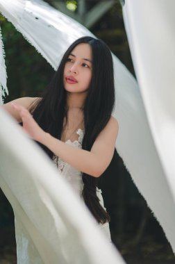 Zarif genç bir kadın, dalgalı beyaz kumaşlar arasında poz verir, dingin güzelliği yakalar. Doğal ışık, bu sakin açık hava ortamında narin özelliklerini geliştirir..