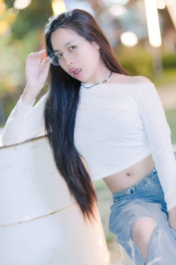 Şık, genç bir kadın, güneş gözlüğü ve günlük kıyafetler takarak, güvenli bir şekilde dışarıda poz veriyor. Uzun saçları aşağıya doğru akıyor, taze ve canlı bir görünüm sergiliyor..