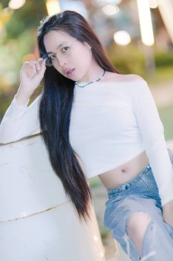 Genç bir kadın beyaz bluzu ve kot kotuyla şık bir şekilde poz veriyor. Uzun saçları, gözlükleri ve sakin ifadesi dışarıda şık bir atmosfer yaratıyor..