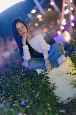 Genç bir kadın canlı bitkiler ve parıldayan ışıklarla çevrili bir şehir bahçesinde düşünceli bir şekilde poz veriyor. Huzurlu atmosfer onun şık görünümünü arttırır..