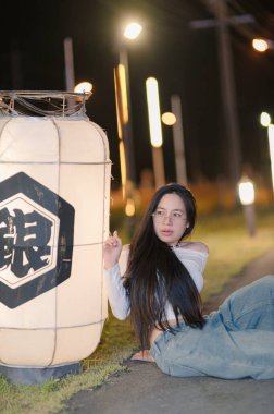 A young woman with long hair poses next to a traditional lantern during a peaceful night. The soft illumination enhances her casual elegance in an urban setting. clipart