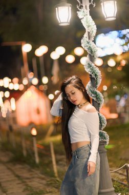 A young woman poses gracefully under a beautifully lit street lamp during a lively nighttime festival. The scene captures her relaxed expression and stylish outfit amidst a backdrop of twinkling lights and festive decorations. clipart