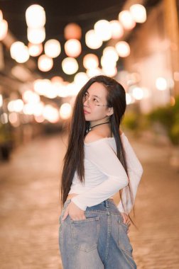 A young woman with long hair poses confidently against a softly lit backdrop. The warm street lights create a serene atmosphere, showcasing her stylish outfit. clipart