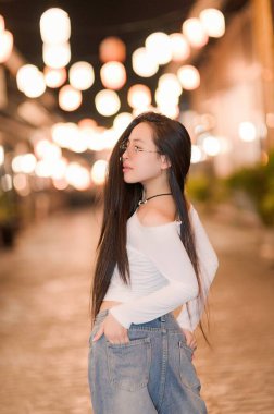 A stylish young woman poses eloquently in a charming urban setting, showcasing her long hair against a backdrop of glowing lanterns, capturing a serene night ambiance. clipart