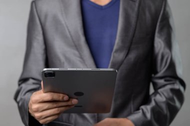 Kendine güvenen, şık gri takım elbiseli bir adam elinde modern teknolojiyi gösteren bir tablet tutuyor. Tarafsız arka plan profesyonelliği ve yeniliği vurguluyor.