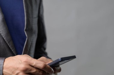 A man in a suit is holding a smartphone with a gray background. The image captures modern technology in a professional yet casual setting, perfect for business-related themes. clipart