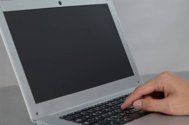 Boş bir ekranla dizüstü bilgisayarın klavyesine yazı yazan bir elin yakın plan görüntüsü, çalışma alanındaki modern teknolojiyi gösteren minimal gri bir arka plan üzerine kurulmuş..