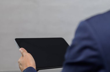 Takım elbiseli bir işadamı gri arka planda boş ekranlı tablet bir cihaz tutuyor. Görüntü modern bir iş ortamı yakalıyor ve teknoloji kullanımını vurguluyor.