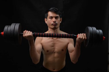 Odaklanmış genç bir adam dambılı güçlü bir pozda kaldırır, kararlılık ve zindeliğe bağlılığını gösterir. Güç ve sağlık gelişimi konuları için ideal.