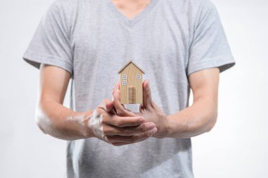 Gri gömlekli şahıs, beyaz bir arka plana karşı ev sahipliği ve gayrimenkul yatırımını sembolize eden küçük ahşap bir ev modeli tutuyor..