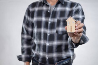 Klasik ekose gömlek giyen bir kişi, ev, tasarım ve gayrimenkul yatırımlarını sembolize eden küçük ahşap bir ev modeli bulundurur..