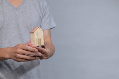 Gri arka planda küçük ahşap bir ev modelini tutan ellerin yakın plan görüntüsü. Bu görsel ev sahipliğinin, konforun ve yaratıcılığın özünü yakalar..
