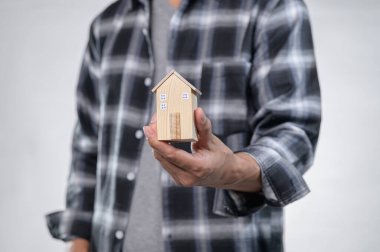 Flanel gömlekli bir kişi ev sahipliği ve emlak yatırımını sembolize eden küçük ahşap bir ev modeli taşıyor. Görüntü, yaratıcılığın ve rüyaların özünü yakalar..