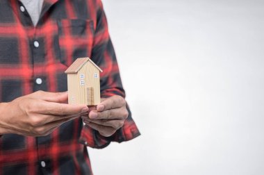 Kırmızı fanila gömlekli bir kişi, yaratıcılığı, konforu ve tasarımda ev ve mimari kavramını sembolize eden küçük ahşap bir ev modeli taşır..