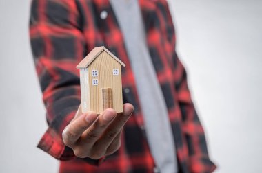 Ekose gömlekli bir kişi, ev sahibi olma ve emlak yatırımı hayallerini sembolize eden el yapımı ahşap bir ev modeli taşıyor..