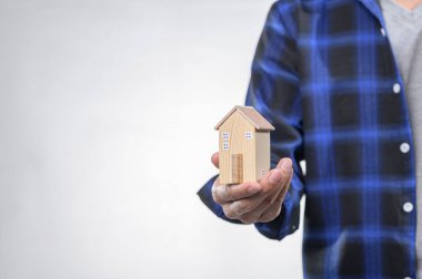 Bir insan bir elinde minyatür ahşap bir ev taşırken sıradan mavi bir kareli gömlek giyer. Bu resim ev, güvenlik ve gayrimenkul yatırımını sembolize ediyor.