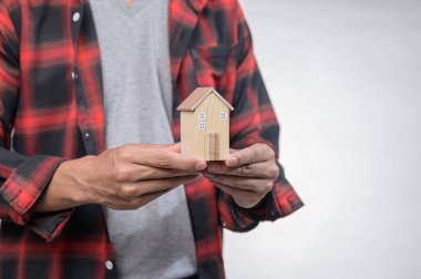 Ekose gömlekli bir kişi, ev sahipliğini ve hayalleri sembolize eden küçük ahşap bir ev modeli taşıyor. Bu resim tasarım, yatırım ve yaşam tarzı seçimlerini temsil eder..