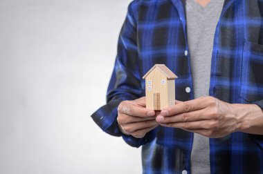 Bir kişi küçük ahşap bir ev modeli tutuyor, ev sahipliğini ve potansiyel yatırımı sembolize ediyor, yumuşak bir arka planda günlük mavi bir fanila gömlek giyiyor,.