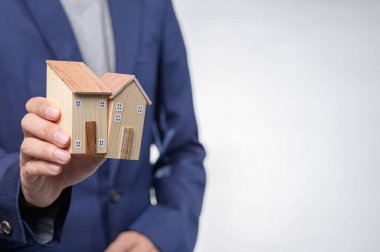Bir iş adamı, emlak piyasasını simgeleyen iki evin ahşap modelini elinde tutuyor. Bu resim yatırım, konut olanakları ve mülkiyet hakkını temsil etmektedir..