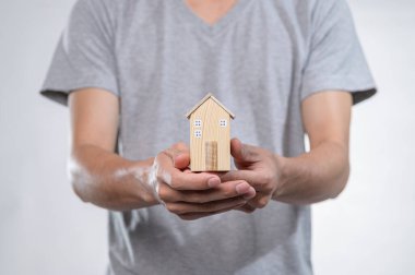 Gri tişörtlü bir kişi iki eliyle minyatür ahşap bir ev modeli sunuyor, ev sahipliği ve yeni başlangıçları simgeliyor, yaratıcılık ve samimiyet anını yakalıyor..